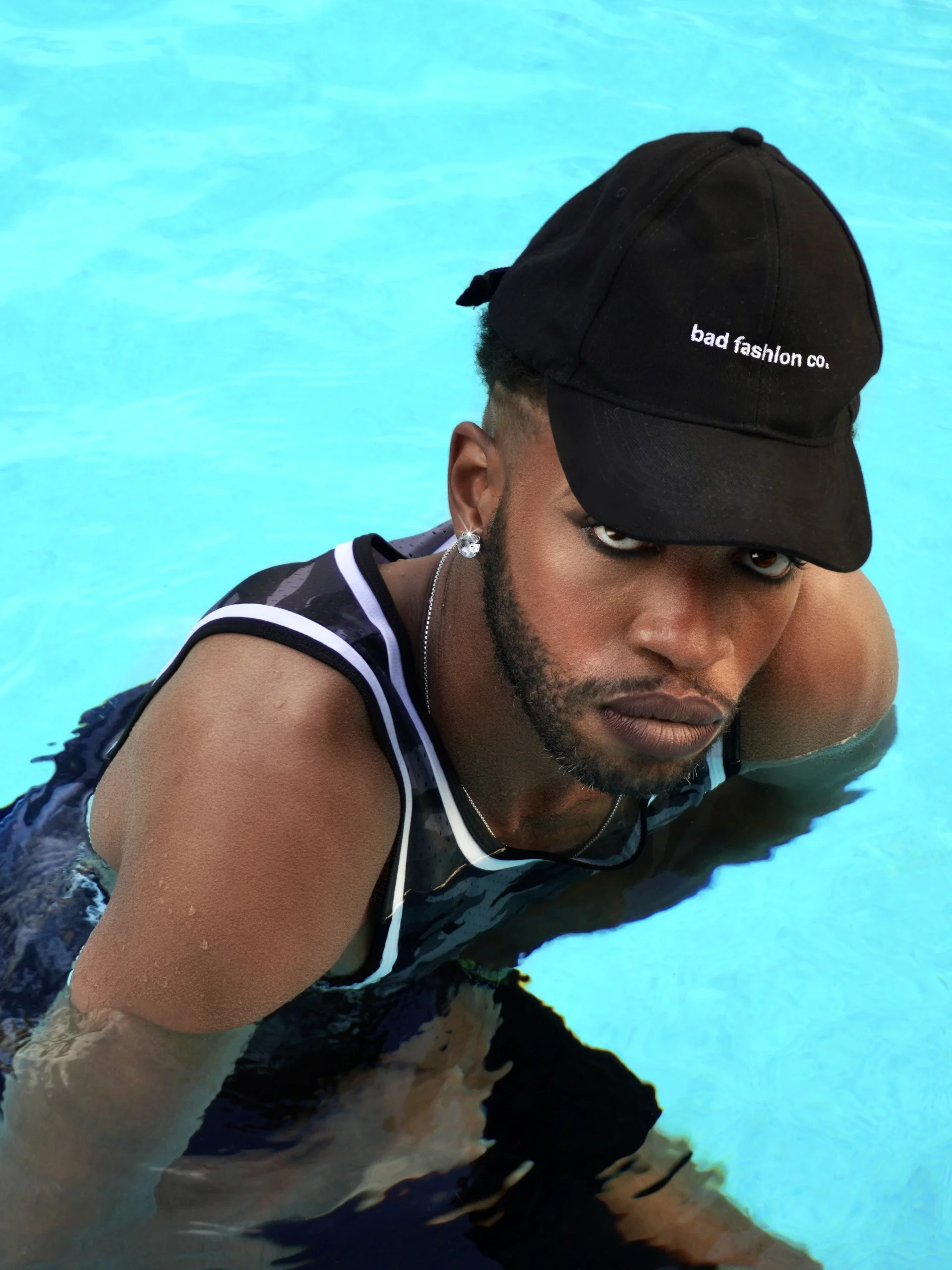 A man in a black &lsquo;bad fashion co.&rsquo; cap looks up at the camera while standing chest-deep in a turquoise swimming pool.